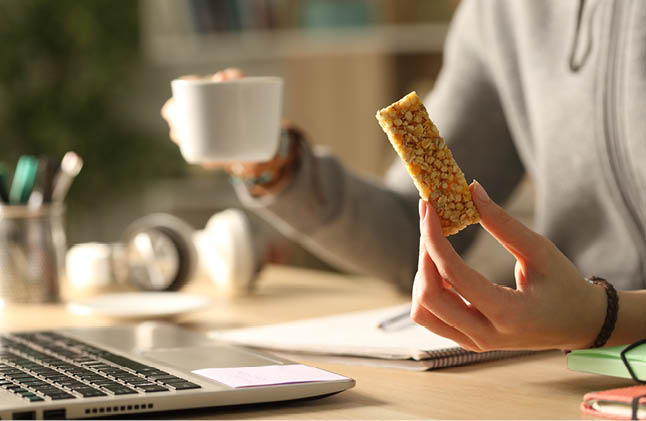 Close up of student girl hands holding coffee cup and cereal snack bar at night studying